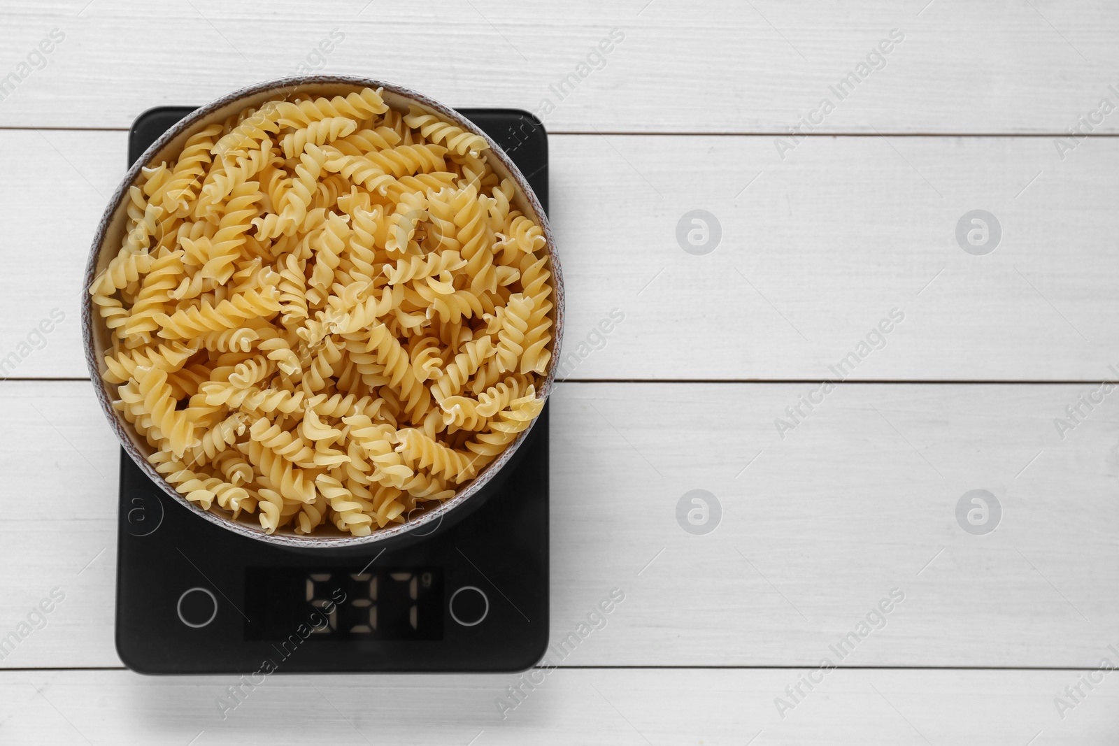 Photo of Electronic scales with uncooked pasta on white wooden table, top view. Space for text