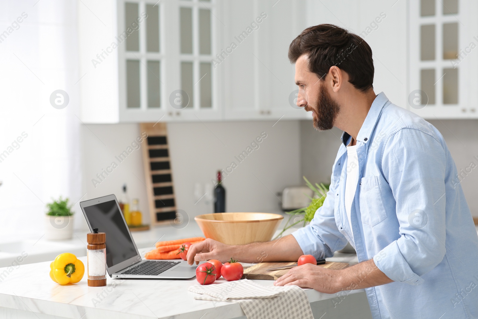 Photo of Man making dinner while watching online cooking course via laptop in kitchen