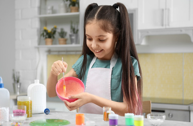Photo of Cute little girl mixing ingredients with silicone spatula at table in kitchen. DIY slime toy