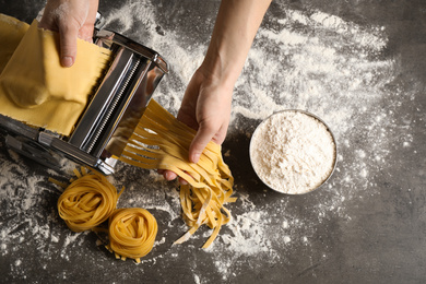 Woman preparing noodles with pasta maker machine at grey table, top view