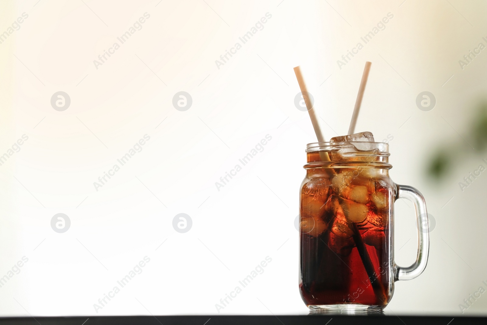 Photo of Mason jar of cola with ice on table against blurred background. Space for text