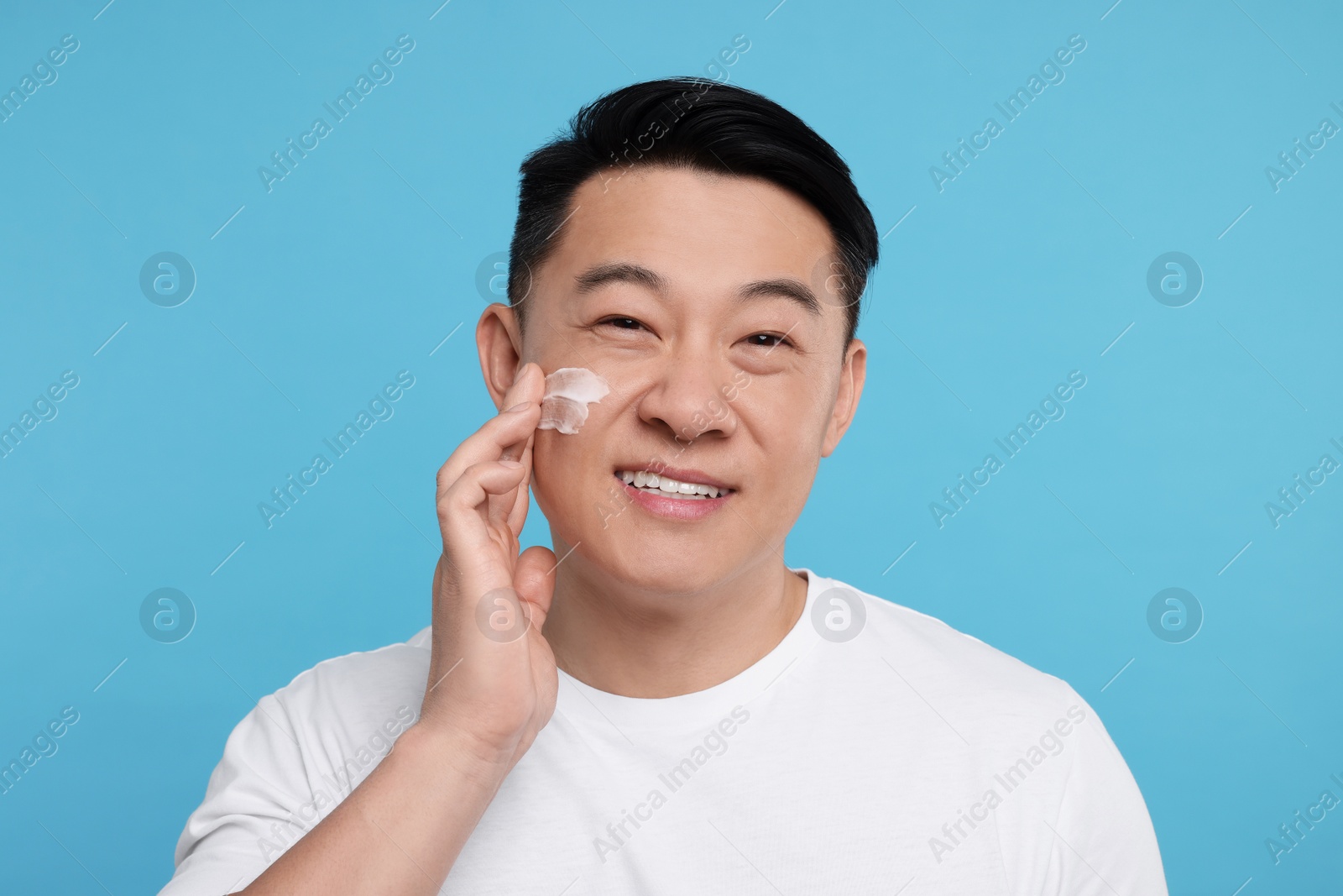 Photo of Happy man applying cream onto his face on light blue background