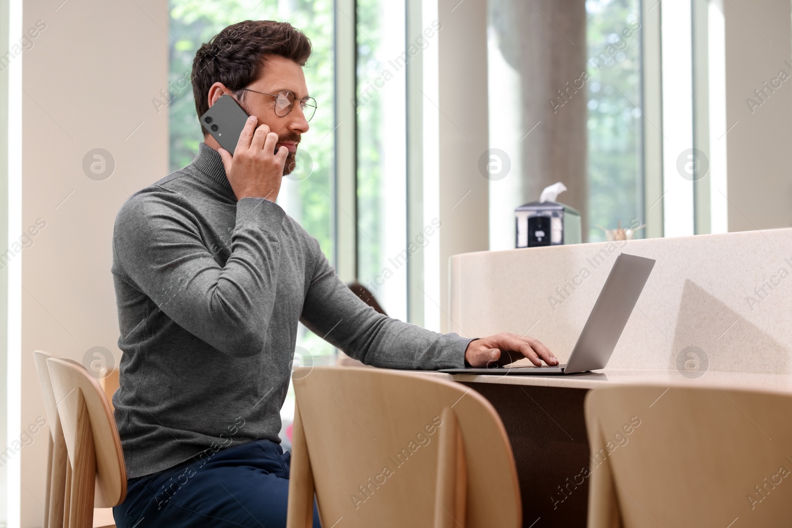 Photo of Handsome man talking on smartphone while using laptop at table in cafe