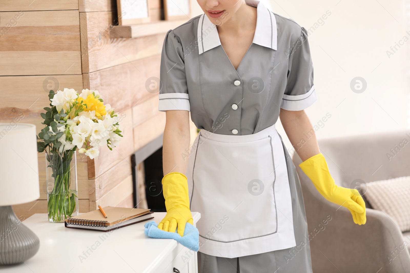 Photo of Young chambermaid wiping dust from furniture in hotel room, closeup