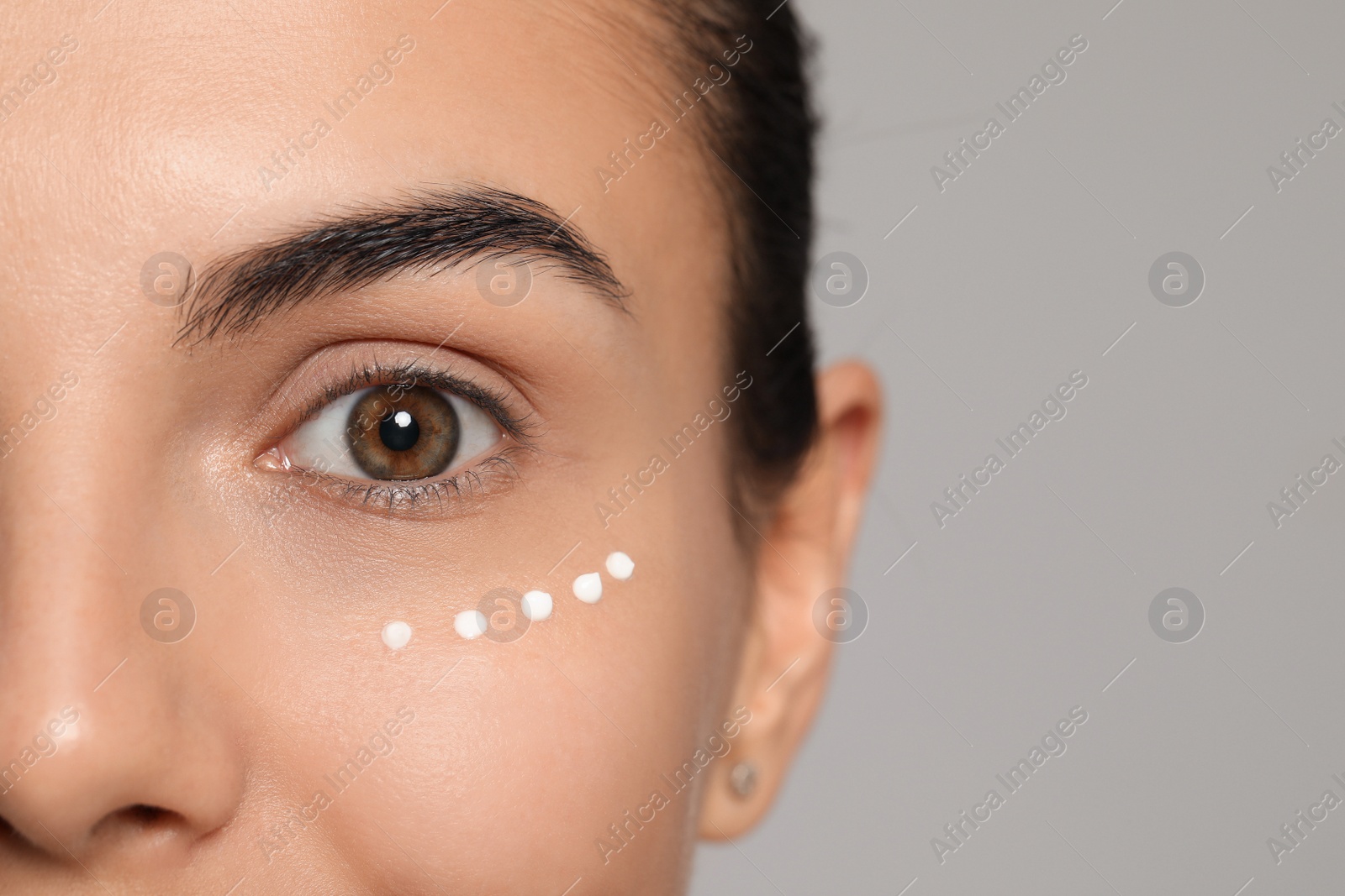 Photo of Young woman with cream on skin under eye against light grey background, closeup. Space for text