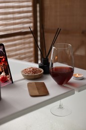 Photo of White wooden tray with glass of wine, comb and burning candle on bathtub in bathroom