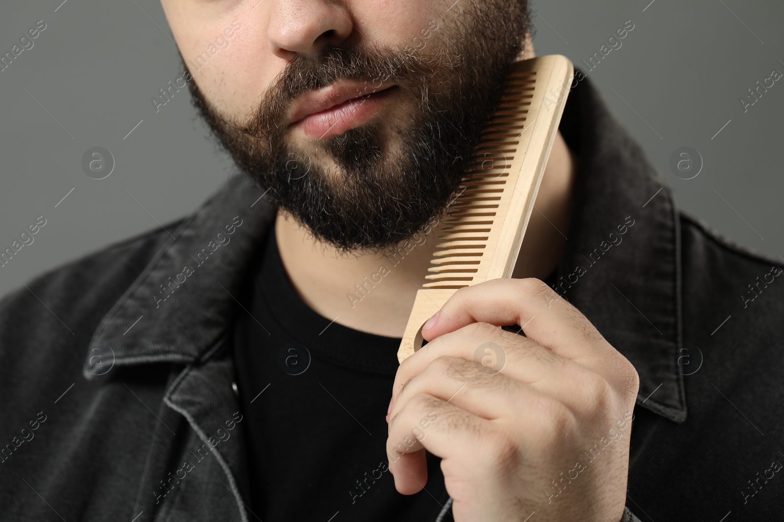 Photo of Handsome young man combing beard on grey background, closeup