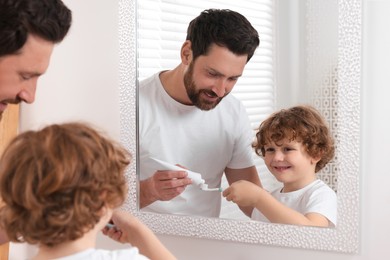 Photo of Father squeezing toothpaste from tube onto son`s toothbrush near mirror in bathroom