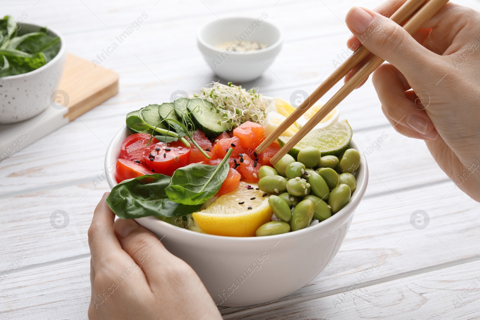 Photo of Woman eating delicious poke bowl with quail eggs, fish and edamame beans at white wooden table, closeup