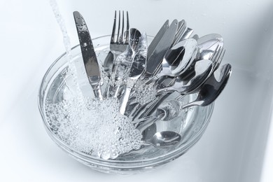 Washing silver spoons, forks and knives under stream of water in kitchen sink