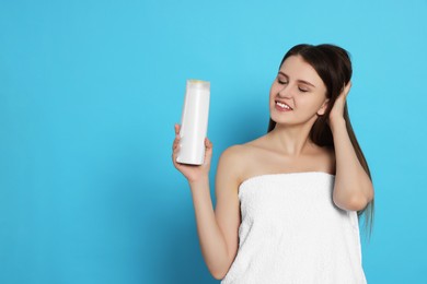 Beautiful young woman wrapped in towel holding bottle of shampoo on light blue background. Space for text
