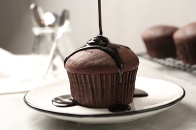 Photo of Pouring chocolate syrup onto delicious fresh cupcake on light grey marble table, closeup