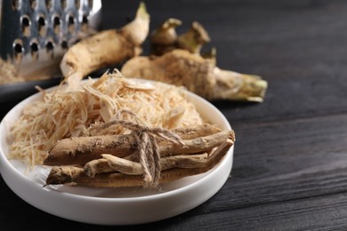 Photo of Plate with whole and grated horseradish roots on black wooden table, closeup. Space for text