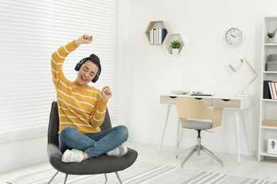 Happy woman in headphones enjoying music and dancing on cosy armchair at home