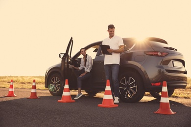 Photo of Instructor with clipboard near car and his student outdoors. Driving school exam