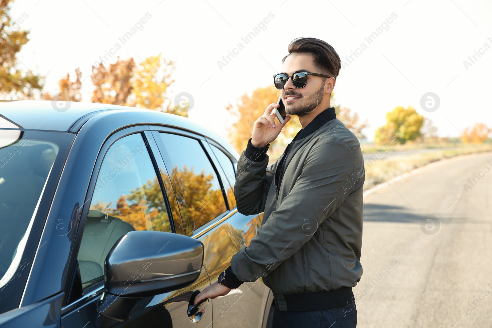 Photo of Young man talking on phone while opening car door, outdoors