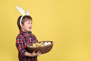 Photo of Cute little boy wearing bunny ears with basket full of dyed Easter eggs on yellow background