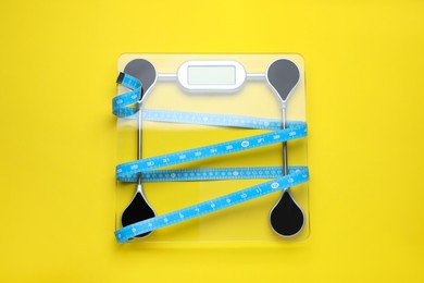 Electronic scales and measuring tape on yellow background, top view