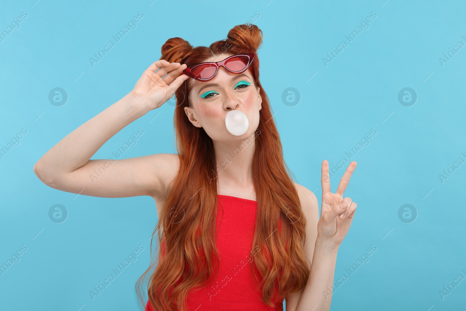 Photo of Portrait of beautiful woman with bright makeup blowing bubble gum and showing peace gesture on light blue background