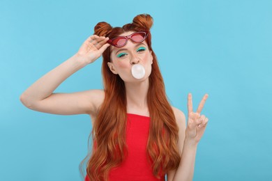 Portrait of beautiful woman with bright makeup blowing bubble gum and showing peace gesture on light blue background