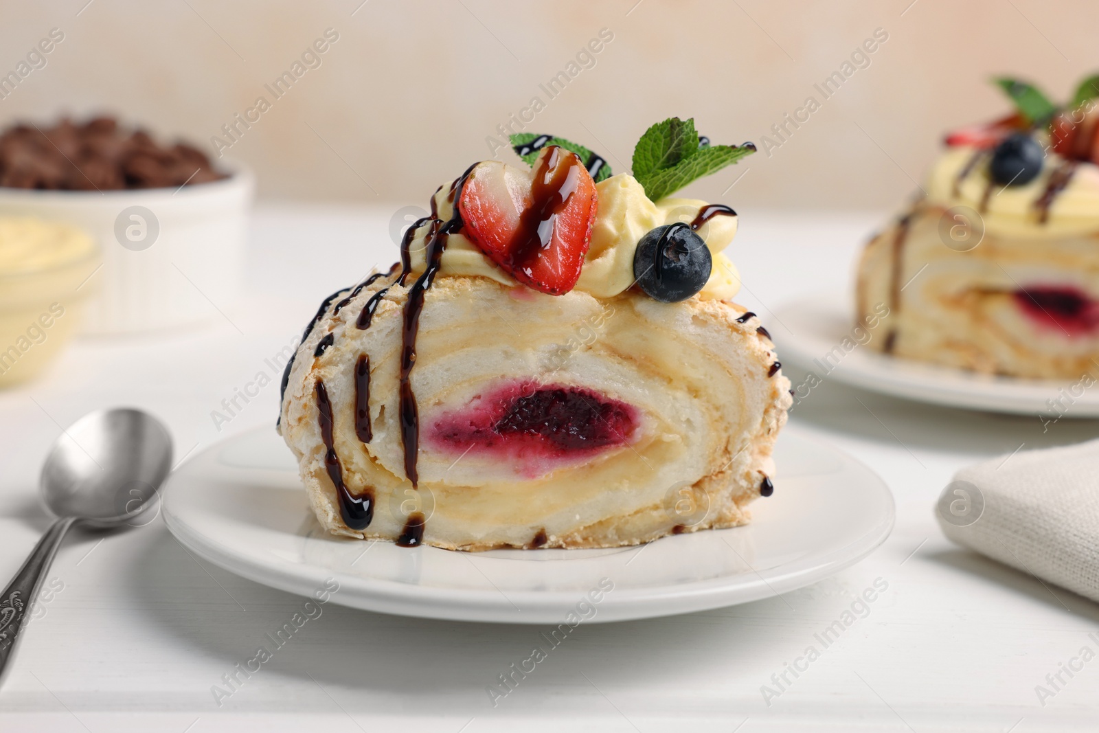 Photo of Piece of tasty meringue roll with jam, cream, strawberry, blueberry and mint on white wooden table, closeup