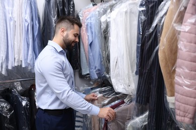 Photo of Dry-cleaning service. Happy worker choosing clothes from rack indoors