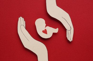 Woman`s health. Hands with newborn paper figure on red background, flat lay
