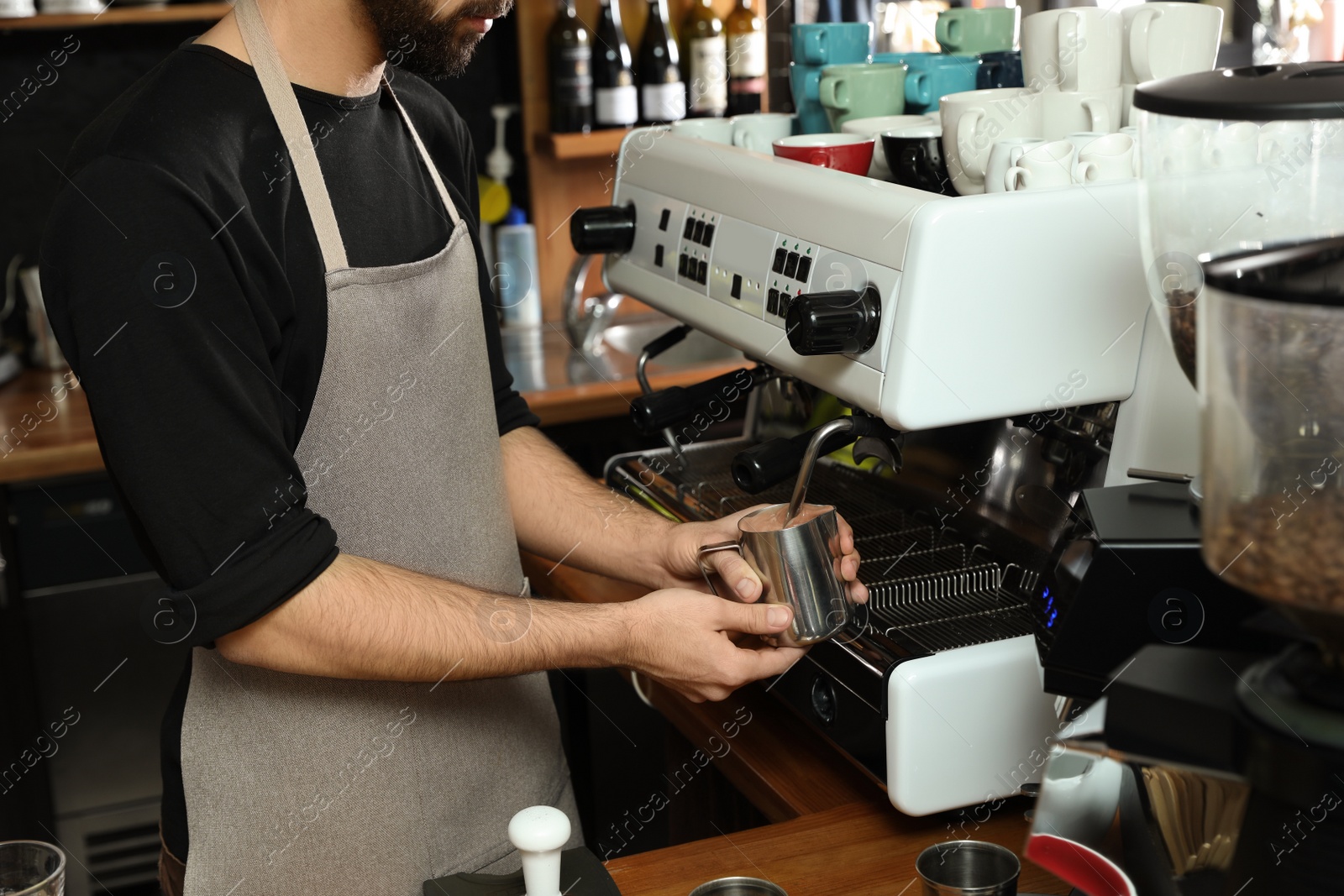 Photo of Barista frothing milk in metal pitcher with coffee machine wand at bar counter, closeup