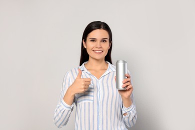 Beautiful young woman holding tin can with beverage and showing thumb up on light grey background