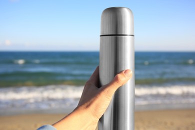 Woman holding metallic thermos with hot drink on beach near sea, closeup