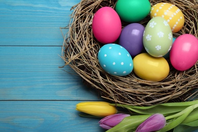 Colorful eggs in nest and tulips on blue wooden background, flat lay. Happy Easter