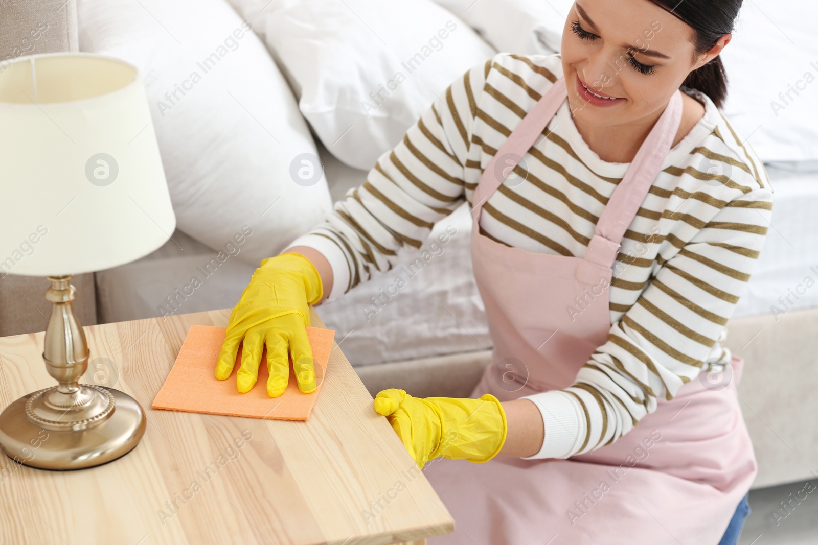 Photo of Young chambermaid wiping dust from nightstand in bedroom, closeup