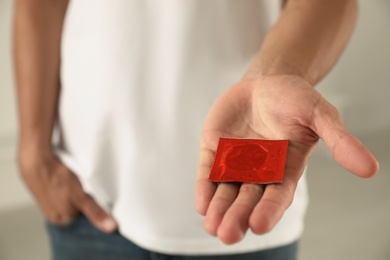 Photo of Young man holding red condom on light background, closeup. Safe sex concept