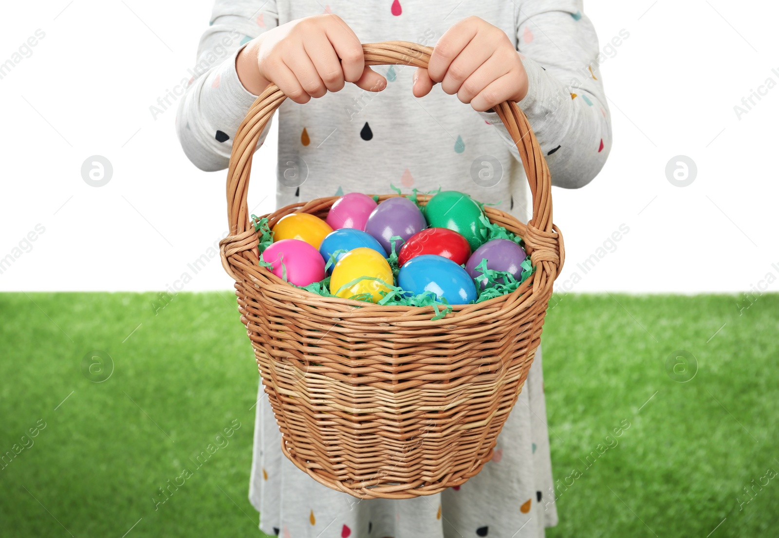 Photo of Little girl with basket full of Easter eggs on green grass against white background, closeup