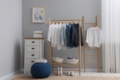 Photo of Wardrobe organization. Rack with different stylish clothes, ottoman and chest of drawers near grey wall in room