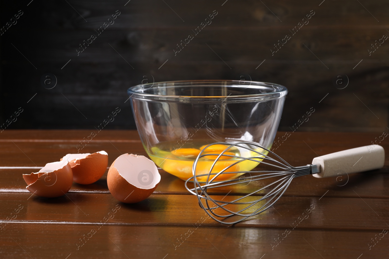 Photo of Metal whisk, raw eggs in bowl and shells on wooden table, closeup