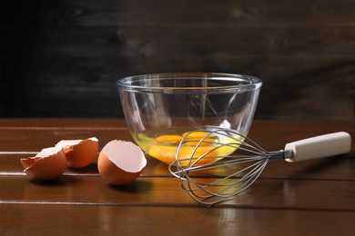 Photo of Metal whisk, raw eggs in bowl and shells on wooden table, closeup