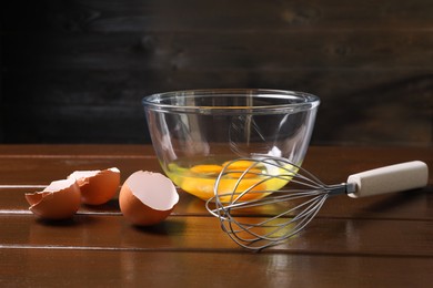 Metal whisk, raw eggs in bowl and shells on wooden table, closeup