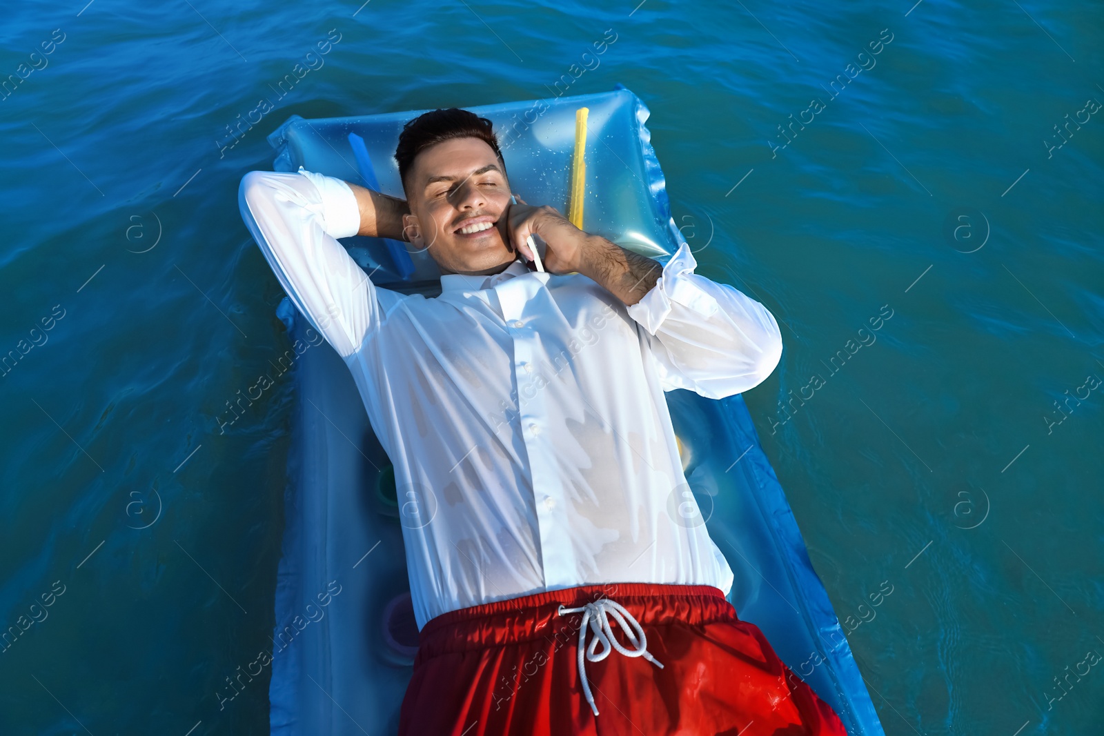 Photo of Happy man talking by mobile phone while floating on mattress in sea. Business trip