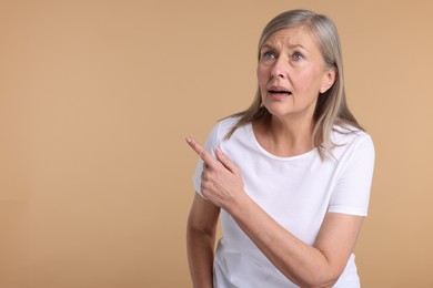 Surprised senior woman pointing at something on beige background, space for text