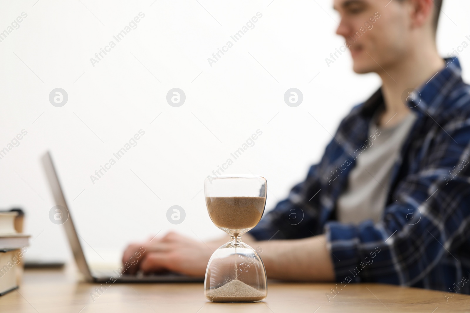 Photo of Hourglass with flowing sand on desk. Man using laptop indoors, selective focus