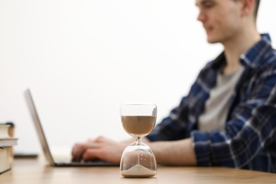 Photo of Hourglass with flowing sand on desk. Man using laptop indoors, selective focus