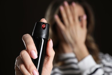 Young woman covering eyes with hand and using pepper spray on black background, focus on canister