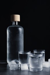 Photo of Bottle of vodka and shot glasses with ice on dark table against black background