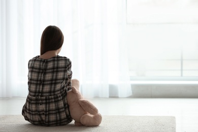 Photo of Upset girl with toy sitting near window indoors. Space for text