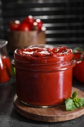 Photo of Jar of tasty tomato paste and basil on grey textured table, closeup