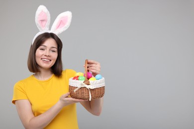 Easter celebration. Happy woman with bunny ears and wicker basket full of painted eggs on grey background, space for text