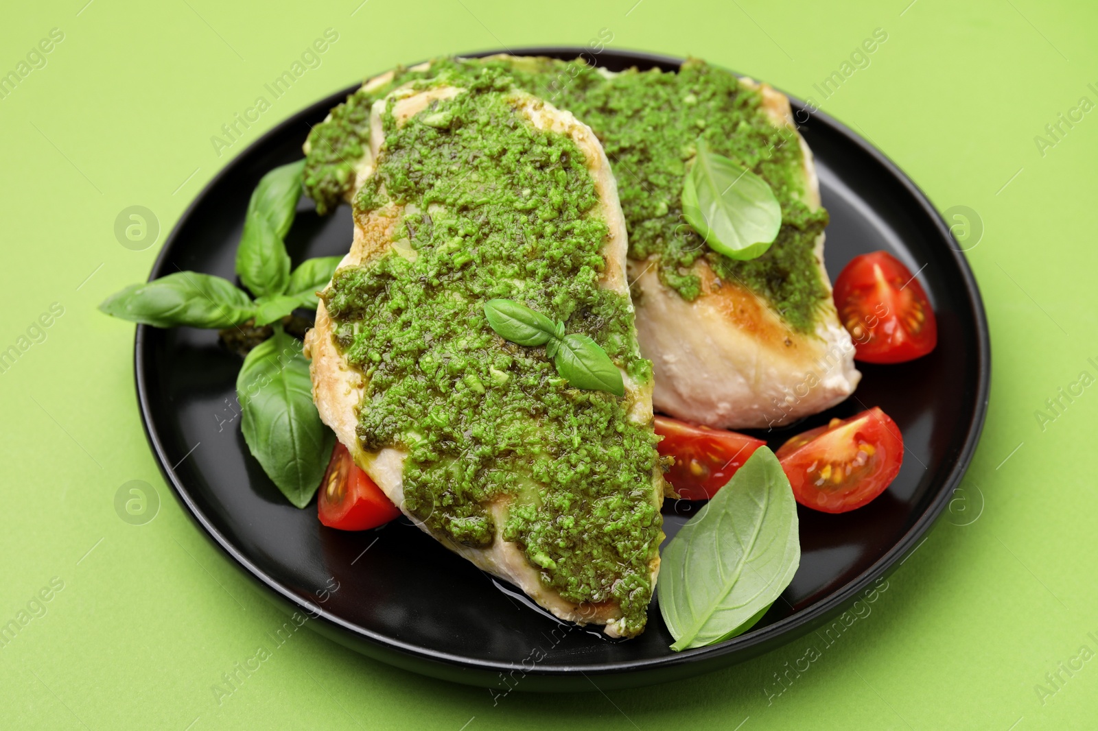 Photo of Delicious chicken breasts with pesto sauce, tomatoes and basil on light green table, closeup