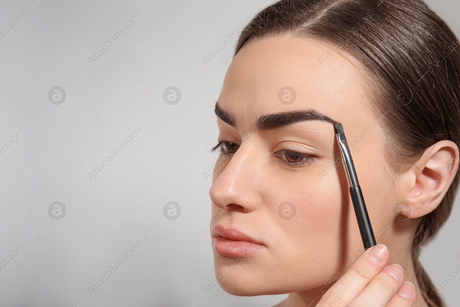 Photo of Young woman correcting shape of eyebrow with brush on light background