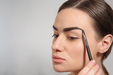 Young woman correcting shape of eyebrow with brush on light background
